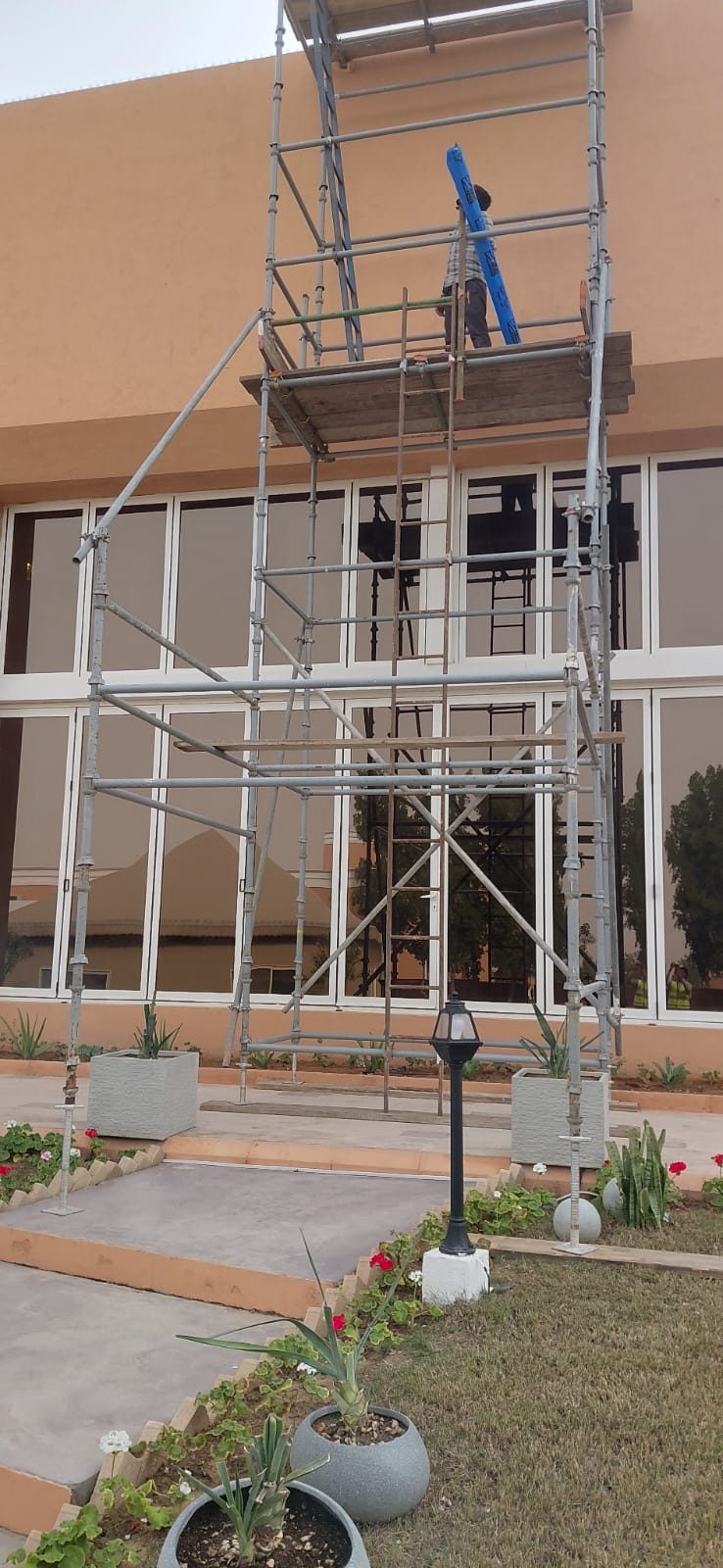 Worker carrying equipment up a tall access scaffold tower erected over a landscaped villa garden