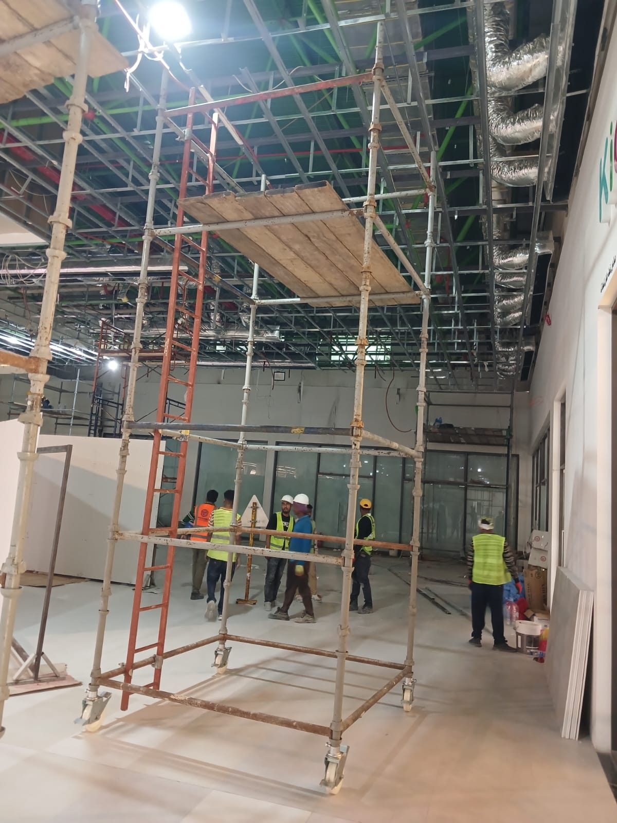 Mobile scaffold towers with timber platforms erected on the floor of an operating shopping mall beneath exposed ceiling services