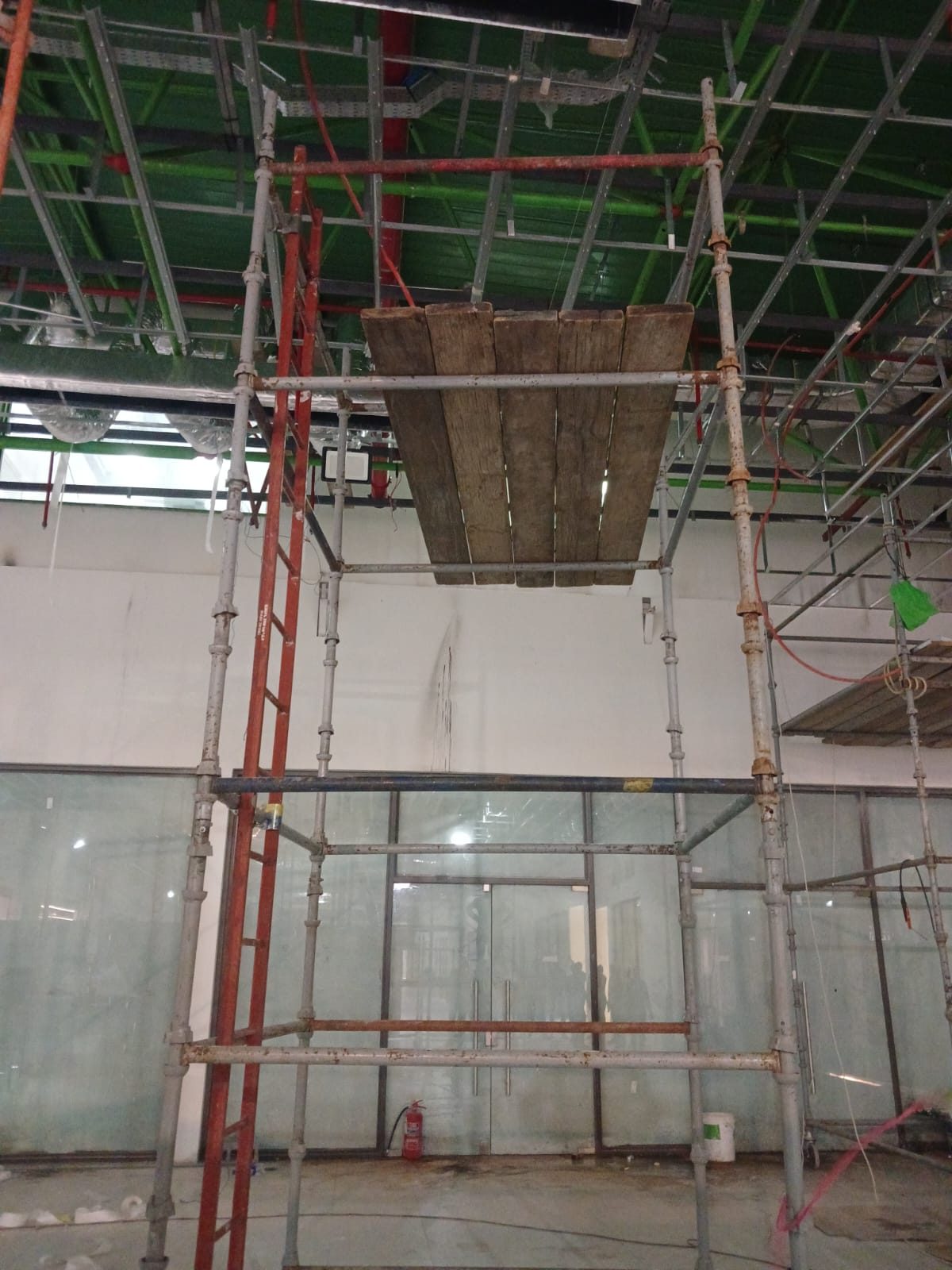 Close view of a boarded scaffold platform and ladder access beneath mall ceiling ductwork and lighting tracks