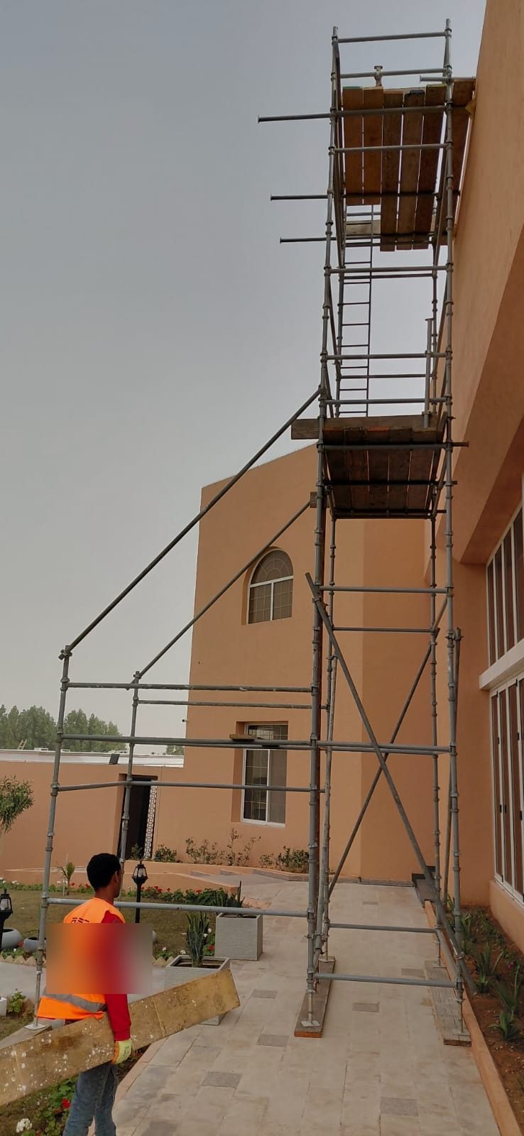 Tall tube-and-clamp scaffold tower with outriggers beside a residential villa, worker in hi-vis vest nearby