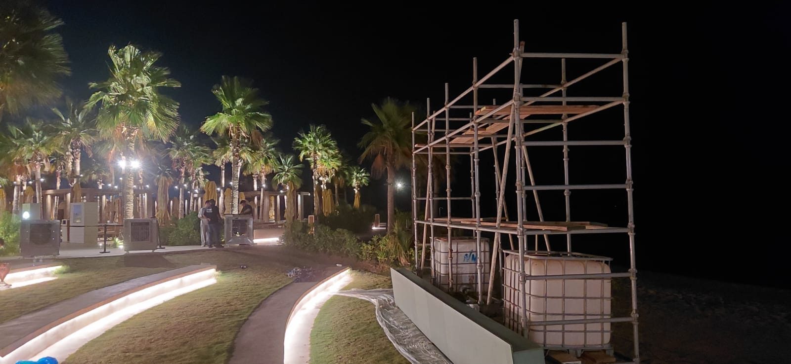 Tube-and-clamp scaffold with timber platforms on a resort waterfront sand area under night lighting