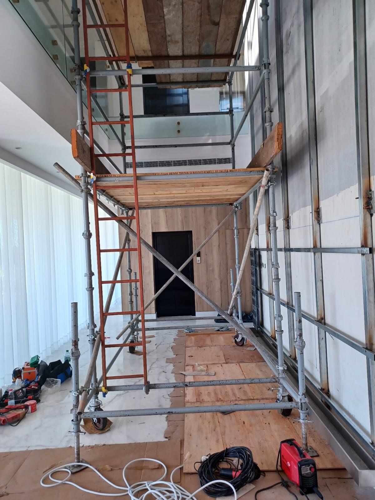 Mobile interior scaffold tower with timber work platform beside a glazed wall and lift lobby