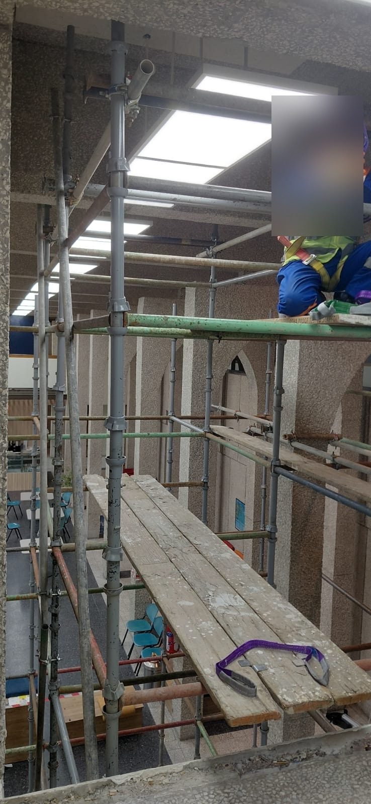 Worker wearing a full fall-arrest harness on an interior scaffold platform beneath skylights