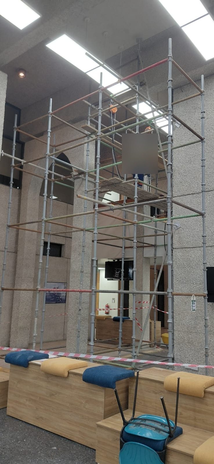 Tube-and-clamp scaffolding inside an institutional atrium with stone columns, an overhead chain hoist and safety barriers